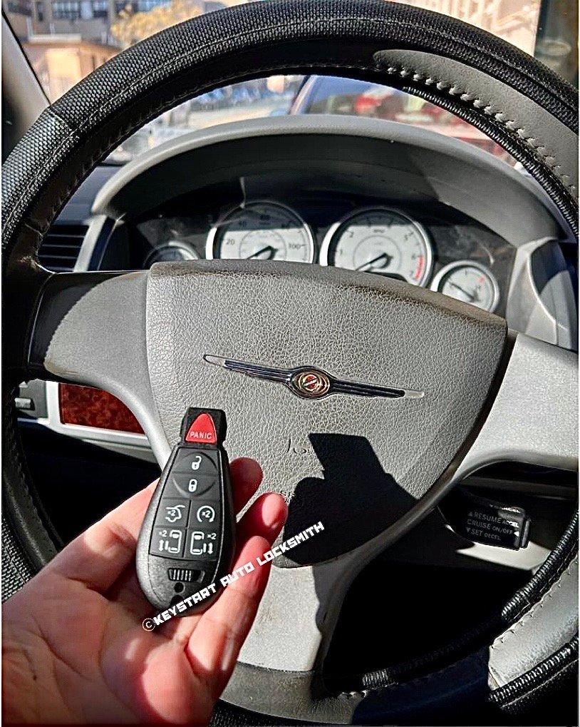 Hand holding a car key fob with a red panic button in front of a Chrysler steering wheel and dashboard gauges in the background.